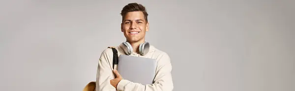 Horizontale Aufnahme eines Studenten in den 20er Jahren mit Laptop und Kopfhörer selbstbewusst stehend auf grauem Hintergrund — Stockfoto