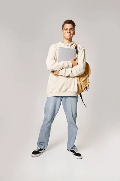 Homem bonito sorridente em seus 20s com laptop confiante em pé contra fundo cinza — Fotografia de Stock