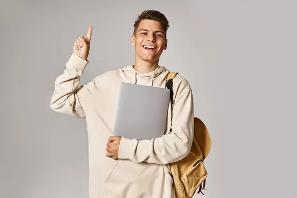 Charming guy in his 20s with laptop and backpack pointing finger to up against grey background — Stock Photo