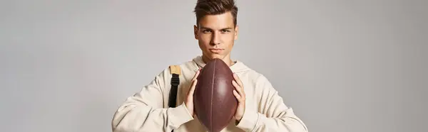 Tiro horizontal de estudiante confiado con la mochila que sostiene la bola de rugby con las manos en el fondo gris - foto de stock