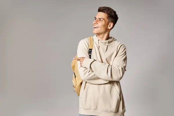 Atractivo joven con mochila y brazos cruzados mostrando el lado con el dedo sobre fondo gris - foto de stock