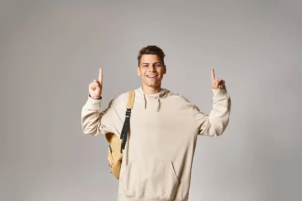Fröhlicher Student in seinen Zwanzigern mit Rucksack, der mit den Fingern nach oben auf grauem Hintergrund zeigt — Stockfoto
