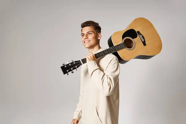Alegre joven con cabello castaño sosteniendo la guitarra en el hombro sobre fondo claro - foto de stock