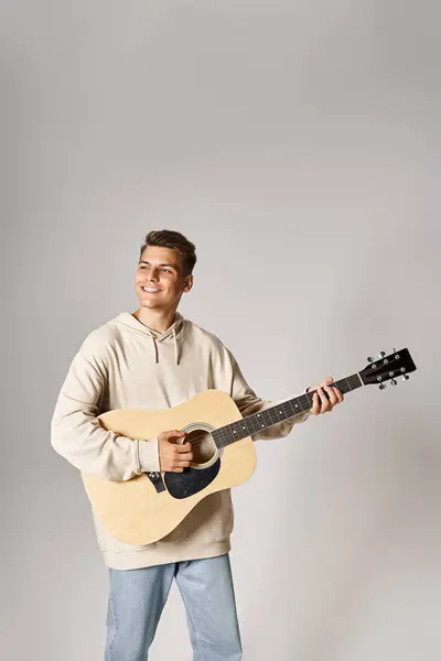 Charming guy in his 20s with brown hair playing on guitar and smiling against light background — Stock Photo