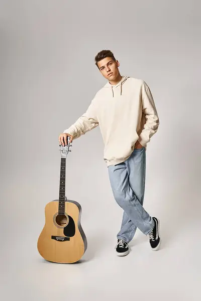 Charismatic young guy in casual outfit with brown hair leaning on guitar against light background — Stock Photo