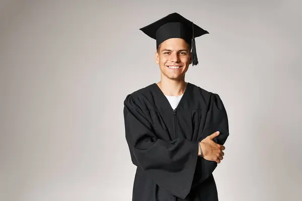 Handsome student in graduate gown and cap with grey eyes folded arms against light background — Stock Photo