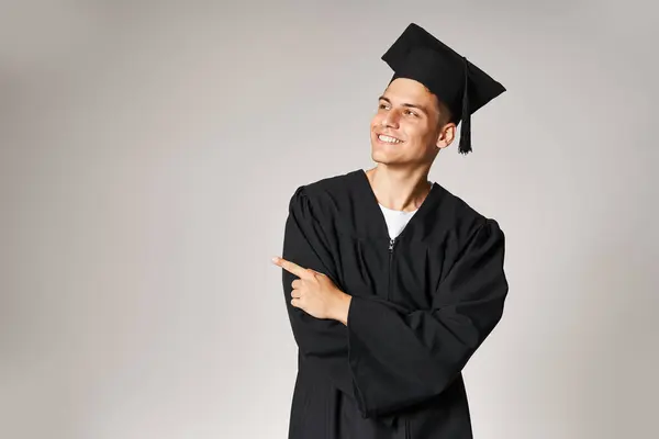 Jovem estudante em vestido de pós-graduação e boné com os braços dobrados apontando dedo no lado em fundo claro — Fotografia de Stock