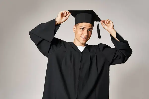Attractive student in gown with grey eyes holding graduate cap on head in light background — Stock Photo