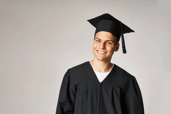 Portrait de jeune étudiant joyeux en robe d'études supérieures et casquette avec les yeux gris sur fond clair — Photo de stock