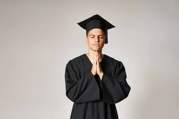 Retrato de esperanzado joven estudiante en vestido de graduado y gorra con las manos dobladas sobre fondo claro - foto de stock