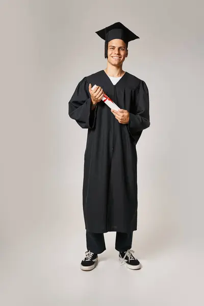 Jovem estudante alegre em vestido de pós-graduação e cap segurando a diploma com as mãos sobre fundo cinza — Fotografia de Stock