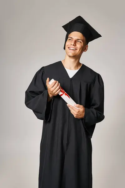 Attractive student in graduate gown and cap holding to diploma with hands and looking to forward — Stock Photo