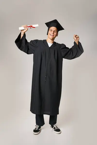 Glücklich schöne Studentin in Diplomkleid und Mütze freut sich über Diplom auf grauem Hintergrund — Stockfoto