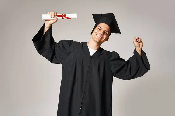 Charming young student in graduate gown and cap rejoices in receiving diploma on grey background — Stock Photo