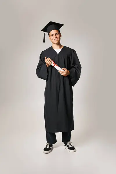Estudante sorrindo em vestido de pós-graduação e cap segurando a diploma com as mãos sobre fundo cinza — Fotografia de Stock