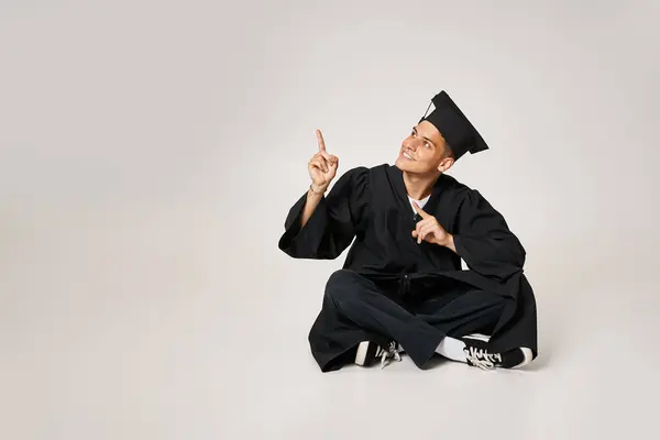 Cheerful young man in graduate gown and cap sitting and showing with fingers to up — Stock Photo