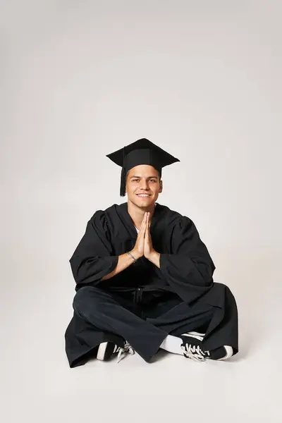 Atractivo joven en vestido de graduado y gorra sentado y plegable manos en fondo gris - foto de stock