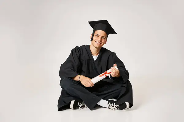 Handsome man in graduate gown and cap sitting and holding to diploma with hands on grey background — Stock Photo