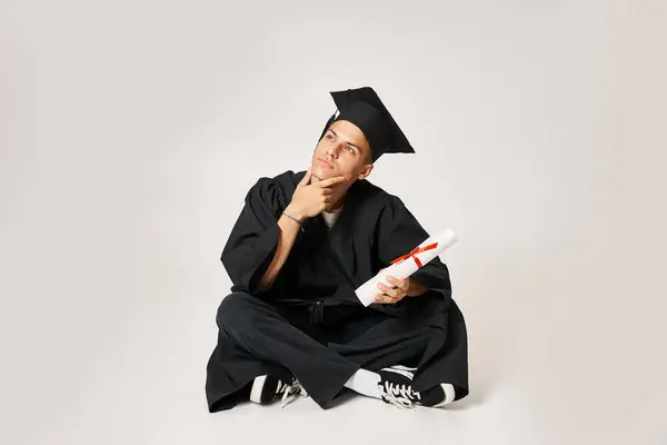 Thoughtful guy in graduate outfit sitting and holding with hand to diploma on grey background — Stock Photo