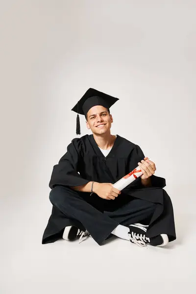 Hombre guapo en vestido de graduado y gorra sentado y sosteniendo el diploma con las manos sobre fondo gris - foto de stock