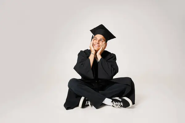Sonriente encantador chico en traje de graduado sentado y sosteniendo con las manos a las mejillas sobre fondo gris - foto de stock