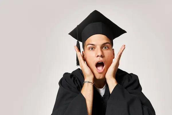 Porträt eines überraschten Studenten mit Diplommütze, der vor grauem Hintergrund in die Kamera blickt — Stockfoto