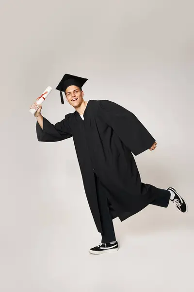 Playful young man in graduate outfit posing with diploma in hand on grey background — Stock Photo