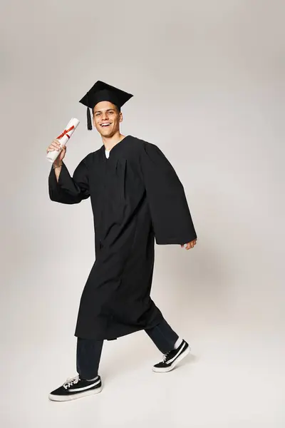 Charming student in graduate outfit walking with diploma in hand on grey background — Stock Photo