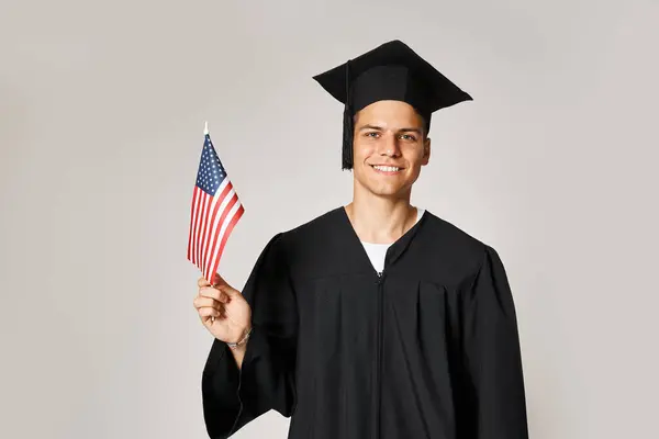 Schöner junger Mann im Diplom-Outfit posiert mit amerikanischer Flagge mit Hand auf grauem Hintergrund — Stockfoto