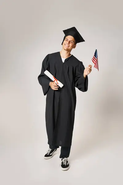 Estudiante feliz en traje de graduado posando con bandera americana y diploma con las manos sobre fondo gris - foto de stock