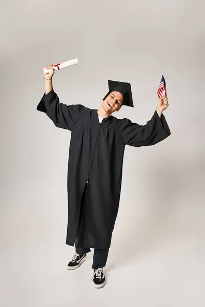 Amerikanischer junger Student im Diplom-Outfit glücklich, sein Studium auf grauem Hintergrund abgeschlossen zu haben — Stockfoto