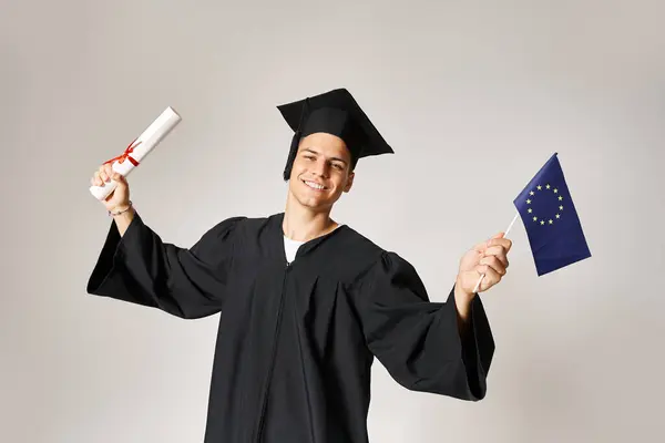 Estudiante europeo en traje de graduado feliz de haber completado sus estudios sobre fondo gris - foto de stock
