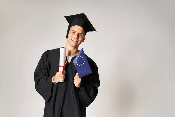 Cara atraente em roupa de pós-graduação posando com diploma e bandeira europeia em mãos em fundo cinza — Fotografia de Stock
