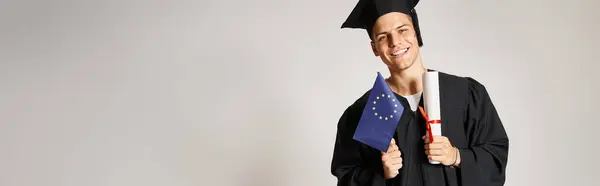 Banner de cara em roupa de pós-graduação posando com diploma e bandeira europeia em mãos em fundo cinza — Fotografia de Stock