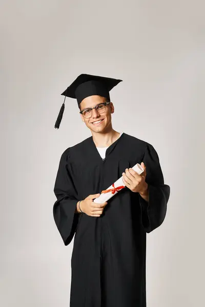 Confident student in graduate outfit with vision glasses smiling with diploma in hands — Stock Photo