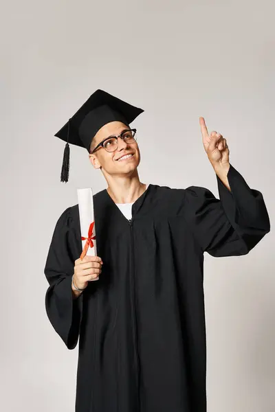 Encantador estudiante en traje de graduado con gafas de visión apuntando con el dedo hacia arriba con diploma en la mano - foto de stock