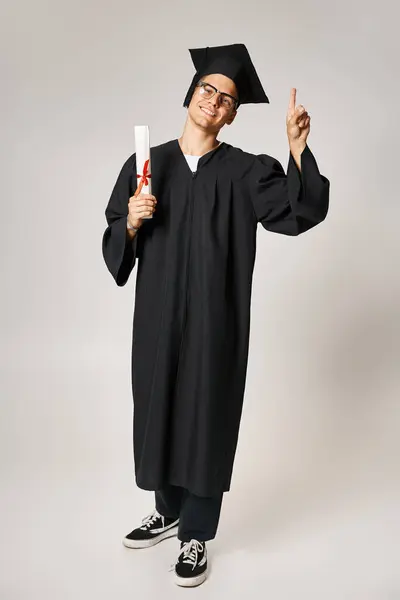 Alegre joven en traje de graduado con gafas de visión apuntando con el dedo hacia arriba con diploma en la mano - foto de stock