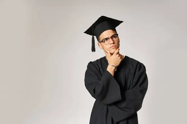 Thoughtful guy in graduate outfit and vision glasses touching hand to jawline on grey background — Stock Photo
