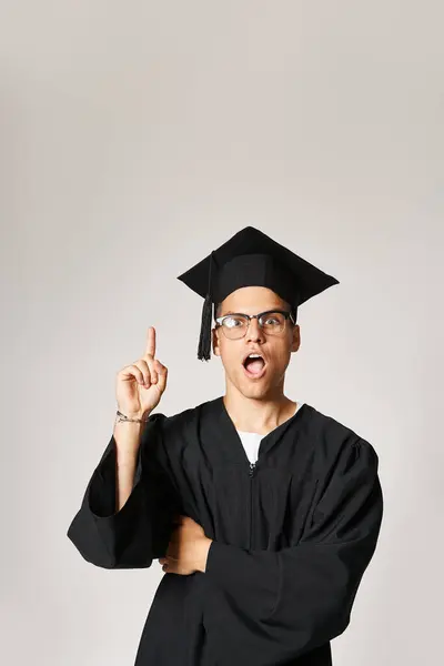 Atractivo joven estudiante en traje de graduado y gafas de visión vienen a la idea sobre fondo gris - foto de stock