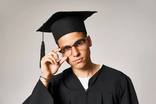 Schöner junger Mann im Diplom-Outfit, der die Hand zur Sehbrille vor grauem Hintergrund berührt — Stockfoto