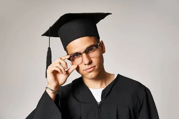Handsome young man in graduate outfit touching hand to vision glasses against grey background — Stock Photo
