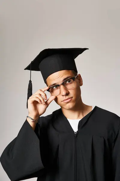 Charming young guy in graduate outfit touching hand to vision glasses and looking to up — Stock Photo