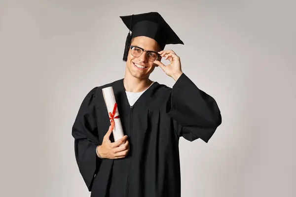 Estudante atraente em roupa de pós-graduação tocando mão para óculos de visão e segurando diploma — Fotografia de Stock