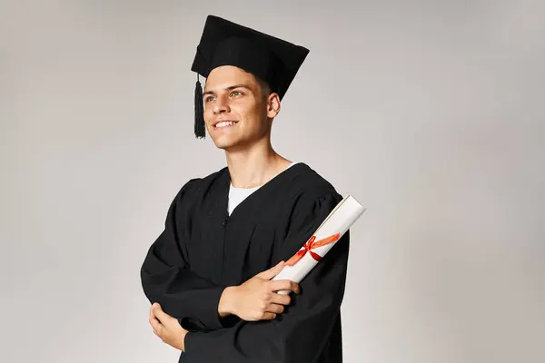 Atractivo joven estudiante en traje de graduado sonriendo y mirando hacia adelante con diploma en la mano - foto de stock