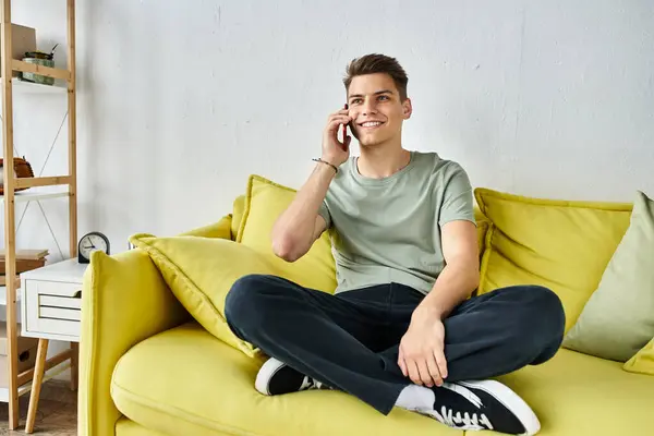 Beau jeune homme aux cheveux bruns à la maison assis sur un canapé jaune et appelant dans le smartphone — Photo de stock