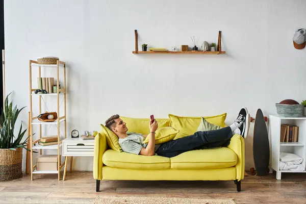 Attractive young man lying on yellow couch in his living room and scrolling to social media — Stock Photo
