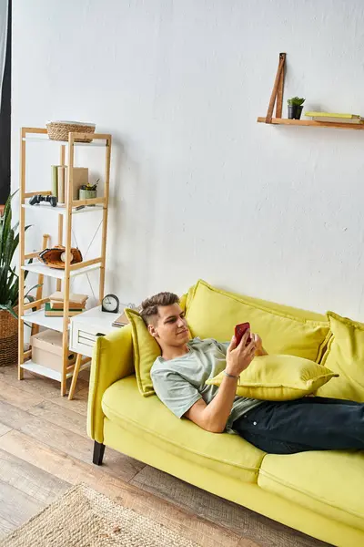 Encantador joven acostado en un sofá amarillo en la sala de estar y desplazándose a las redes sociales - foto de stock