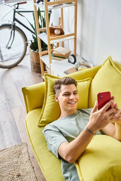 Улыбающийся молодой студент лежит на желтом диване в гостиной и прокручивается в социальных сетях — стоковое фото