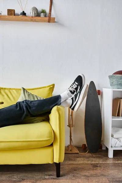 Tiro recortado de las piernas del hombre en zapatillas negras acostado para cruzar en el sofá amarillo en la sala de estar - foto de stock