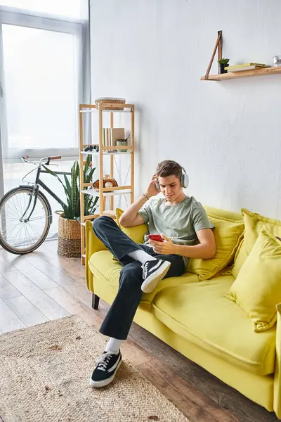 Handsome young man in headphones sitting on yellow couch at home and scrolling to social media — Stock Photo